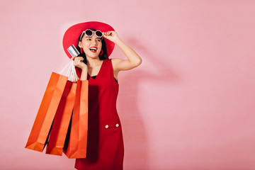 Portrait of a happy Asian pretty girl holding shopping bags while Hold Credit Cards and sunglasses looking away isolated over pink background,colorful shopping concept,red.