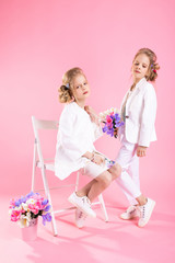 Twins girls in light clothes with bouquets of flowers posing near a chair on a pink background.