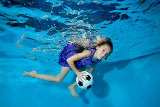 A little girl with a football ball in her hands engaged in sports and swims underwater in the pool in a purple swimsuit. Portrait. Underwater photography. The horizontal orientation of the image