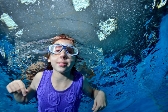 A little girl in swimming glasses is swimming underwater in the pool on a blue background and looking at me. Portrait. Close up. Bottom view. Underwater photography. Horizontal view of the image
