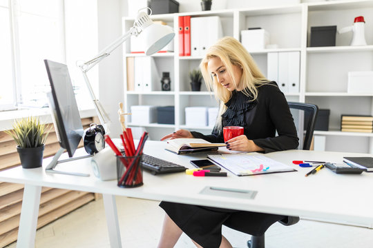 A young girl is sitting at a table in the office, holding a cup and reading a book.