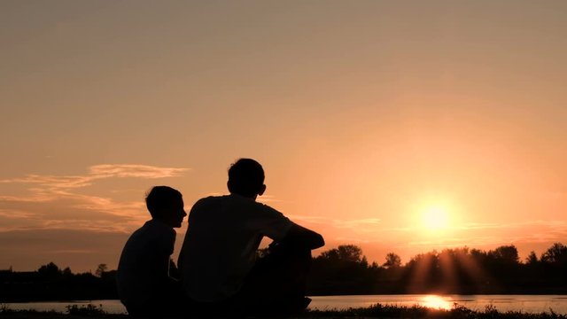 Silhouette of two people communicating on the bank of a river or lake. Father and son are sitting on the waterfront and watch the sunrise or sunset.