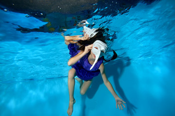 Fototapeta na wymiar A little girl in a masquerade white mask with a long nose swims underwater in the pool on a blue background in a purple dress and looks at the camera. Shooting underwater. Horizontal orientation
