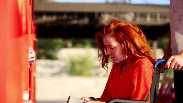 Young Man Rolls His Girlfriend In A Wheelchair To The Bookshelf, She Takes Her Favorite Book And Tells Him About This Book, Outdoors