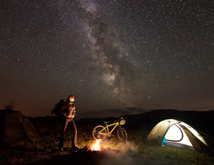 Young woman cyclist having a rest at night camping near burning campfire, illuminated tourist tent, mountain bike under incredible beautiful evening sky full of stars and Milky way. Tourism concept