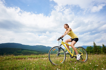 Athlete woman cyclist riding on yellow mountain bicycle on a grassy hill, enjoying summer day in the mountains. Outdoor sport activity, lifestyle concept