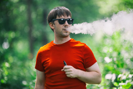 A young white bearded man in red t-shirt and in sunglasses is vaping an electronic cigarette in the forest in the summer. Close up.