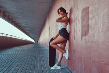 Portrait of a beautiful girl dressed in shorts and t-shirt posing with skateboard while leaning on a wall at the sidewalk under the bridge. © Fxquadro