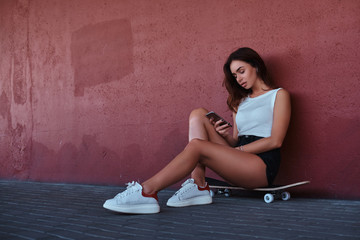 Portrait of a sensual hipster sitting on a skateboard using a smartphone and listening to music while leaning on the wall.