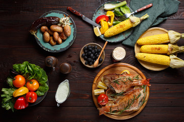 Dinner table with fried fish, corn, salad, grilled potatoes and eggplant, paprika, olives, tomato, celery and sauce, beer top view.
