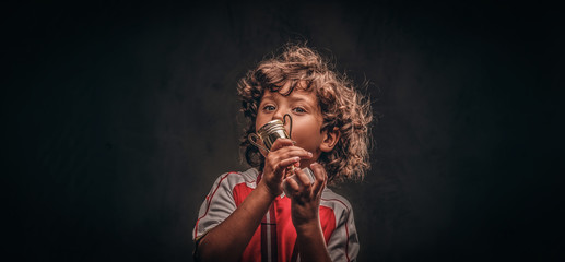 Cute little champion boy in sportswear with gold medal kissing the winner's cup. Isolated on the dark textured background.