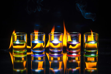 Five multicolored full of drinks shot glasses reflected on the glass surface behind which the flame burns on a black background.