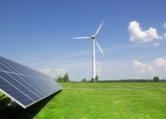 wind generator of electricity from three blades and solar panels of a battery of photocells against a background of clouds and a blue sky green grass