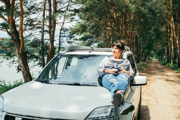 young pretty woman sitting on car hood drinking coffee. lake with forest on background