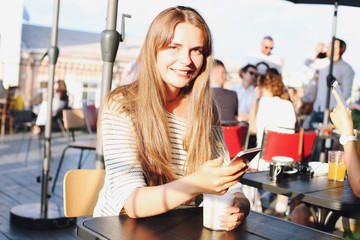 Beautiful young woman sitting in hipster coffee shop on roof top holding mobile cell phone, drinking cappuccino beverage. Attractive female at terrace cafe in Europe. Background, close up, copy space.