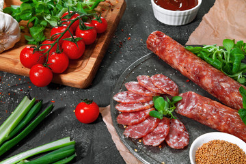 Sliced sausage on the board on dark background. Sausage with spices, pepper, spinach, cherry tomatoes. View at an angle of 45 degrees. Place for text. Advertising of sausage