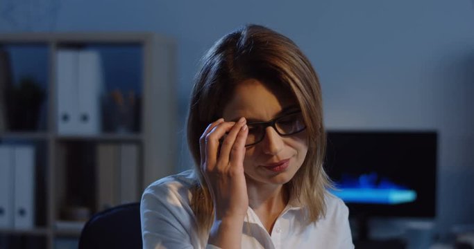 Close up of the Caucasian young pretty woman in the white blouse and glasses working at the computer in the office at night and smiling, then taking oof glasses. Inside