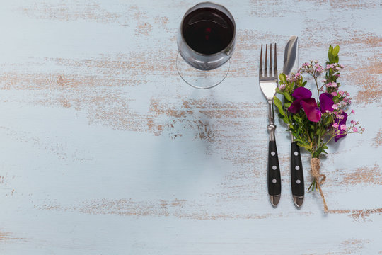 Rustic table setting with purple flowers and glass of red wine on light wooden table.