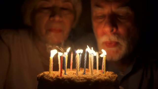 Loving senior couple celebrating anniversary with cake at home in the evening. Happy elderly family hugging, cuddling together, make wishes and blowing out candles. Focus on the cake