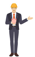 Businessman in construction helmet holding a piggy bank and showing something beside of him
