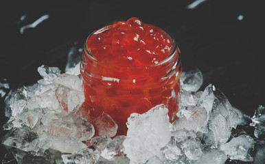 Delicious red caviar in a glass jar on crushed ice on black stone background, selective focus and...
