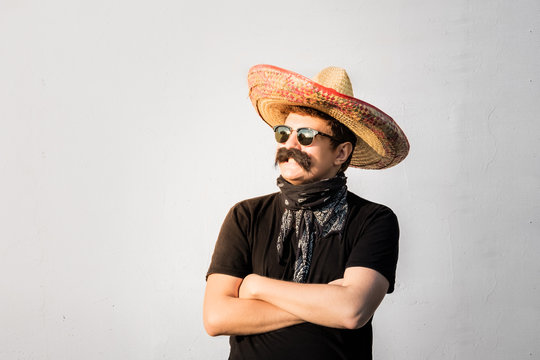 Young Male Person Dressed Up In Traditional Mexican Sombrero, False Moustache, Bandana And Sunglasses. Movember Or Halloween Concept Of Man Posing As Bandit Or Western Style Gangster