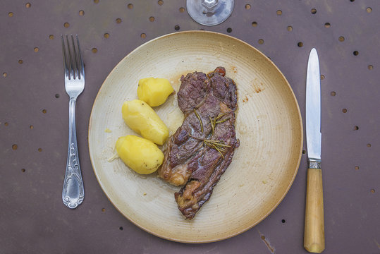 Grilled beafsteak with potatoes on rustic plate, glass and cutlery on dark metallic background top view