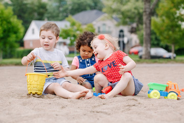 Portrait of three cute Caucasian and hispanic latin toddlers babies children sitting in sandbox playing with plastic colorful toys. Little girls and boy friends having fun together on playground. - obrazy, fototapety, plakaty
