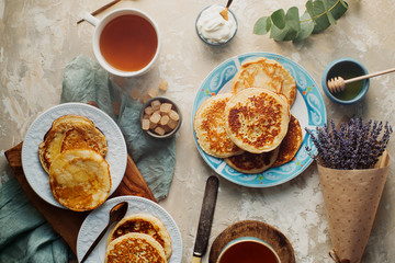 Tea, homemade pancakes, honey, cream sauce and lavender on light concrete table, romantic breakfast pancake party, various morning food, healthy lifestyle. Selective focus, top view