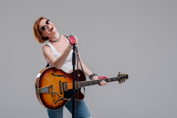 Portrait of a young woman musician with an electric guitar in her hands and studio microphone on a gray background. She wears black sunglasses and plays rock and roll loudly
