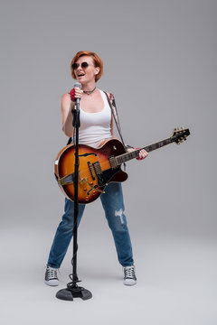 Young woman musician with an acoustic guitar in hand on a gray background. He laughs and plays rock and roll loudly. Full-length portrait