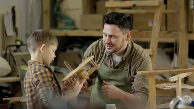 Medium shot of professional male carpenter in apron inspecting wooden airplane, then gifting it to his little son while spending time with him in workshop