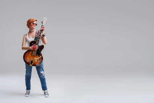 Young woman musician with an acoustic guitar in hand on a gray background. He laughs and plays rock and roll loudly. Full-length portrait. On the right there is space for text