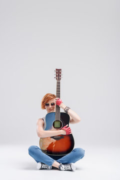 Portrait of a young woman with an acoustic guitar in hands on a gray background. Sits on the floor and hugs a musical instrument