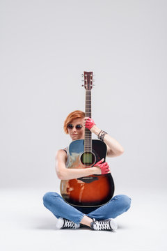 Portrait of a young woman with an acoustic guitar in hands on a gray background. Sits on the floor and hugs a musical instrument