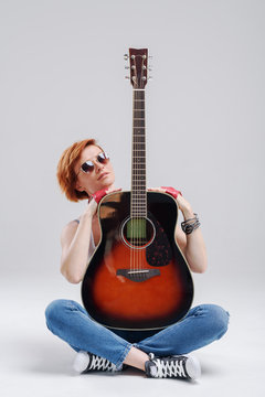 Portrait of a young woman with an acoustic guitar in hands on a gray background. Sits on the floor and hugs a musical instrument