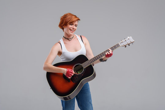 Young woman musician with an acoustic   guitar in hand  on a gray background. He laughs and plays rock and roll loudly. 