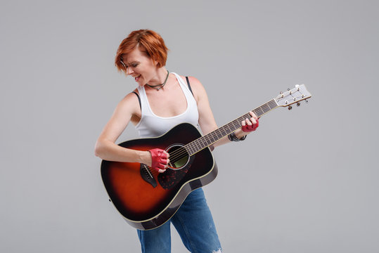Young woman musician with an acoustic   guitar in hand  on a gray background. He laughs and plays rock and roll loudly. 