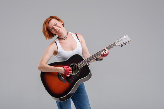 Young woman musician with an acoustic   guitar in hand  on a gray background. He laughs and plays rock and roll loudly. 