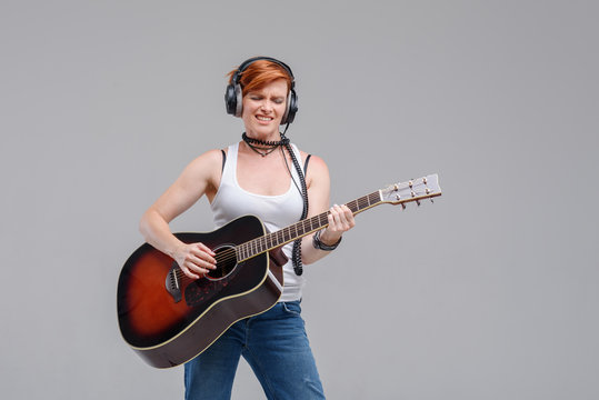 Young woman musician with an acoustic   guitar in hand and in headphones on a gray background. He laughs and plays rock and roll loudly. 