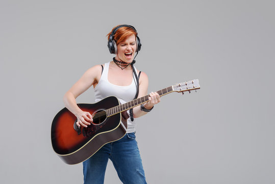 Young woman musician with an acoustic   guitar in hand and in headphones on a gray background. He laughs and plays rock and roll loudly. 