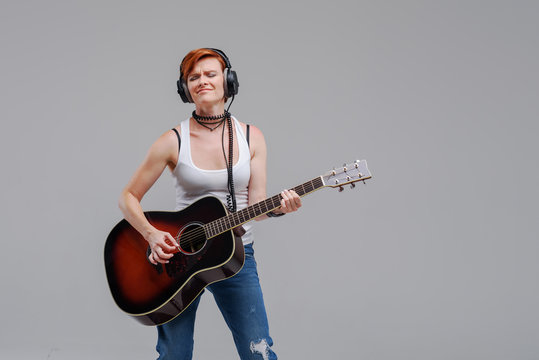 Young woman musician with an acoustic   guitar in hand and in headphones on a gray background. He laughs and plays rock and roll loudly. 
