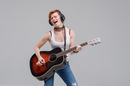 Young woman musician with an acoustic   guitar in hand and in headphones on a gray background. He laughs and plays rock and roll loudly. 
