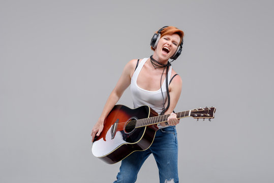 Young woman musician with an acoustic   guitar in hand and in headphones on a gray background. He laughs and plays rock and roll loudly. 