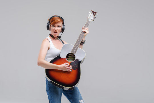 Young woman musician with an acoustic   guitar in hand and in headphones on a gray background. He laughs and plays rock and roll loudly. 