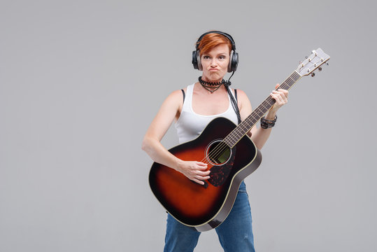 Young woman musician with an acoustic   guitar in hand and in headphones on a gray background. He laughs and plays rock and roll loudly. 