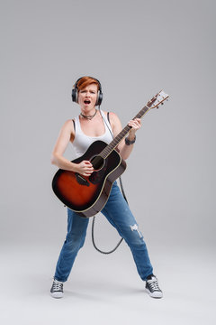 Young woman musician with an acoustic   guitar in hand and in headphones on a gray background. He laughs and plays rock and roll loudly. Full-length portrait