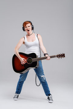 Young woman musician with an acoustic   guitar in hand and in headphones on a gray background. He laughs and plays rock and roll loudly. Full-length portrait