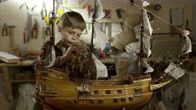 PAN of concentrated little boy applying paint coat on ship model standing on workbench in garage