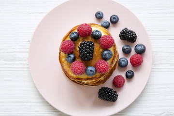 Pancakes with berries on a pink plate over white wooden table, top view.